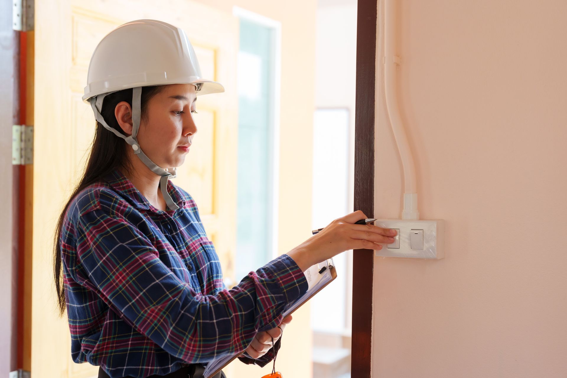 Electrician in a hard hat is inspecting an electrical outlet with a clipboard in hand