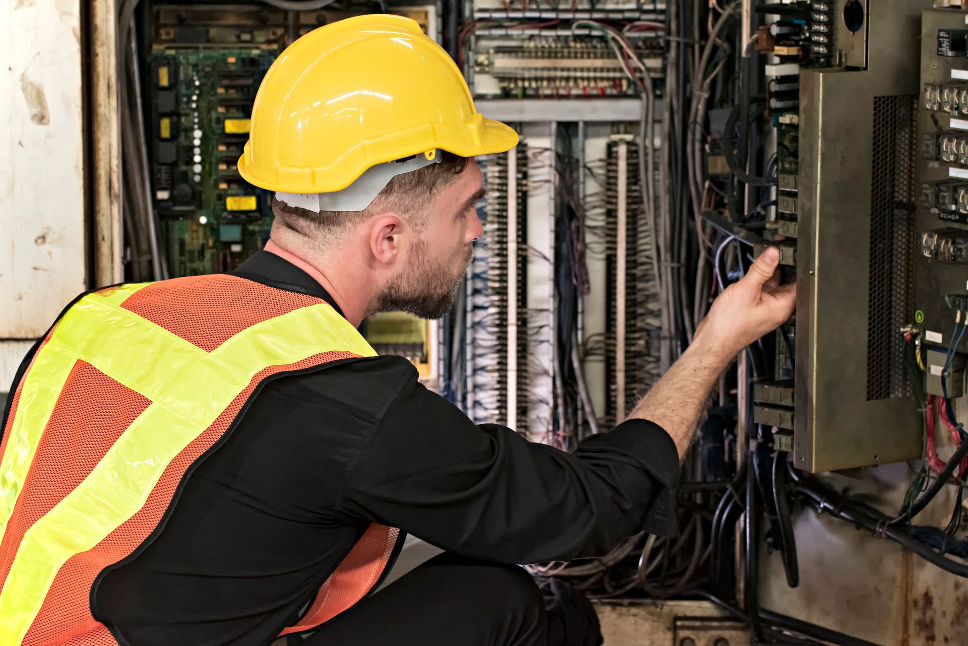An electrician in a yellow hard hat and safety vest is examining electrical wiring