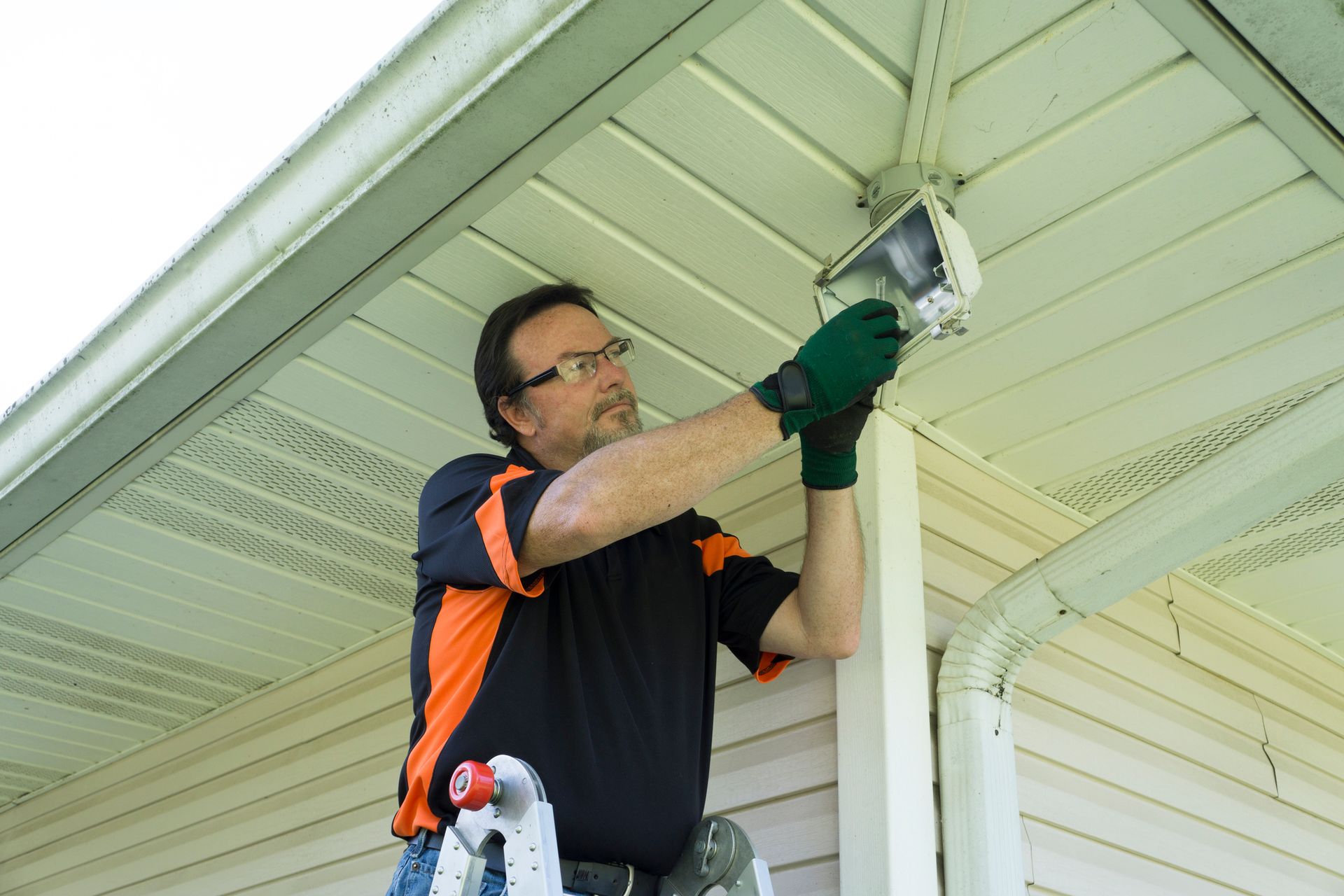 A man on a ladder is installing a security light on a house's exterior corner