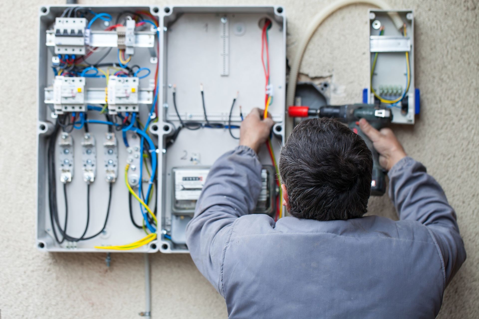 An electrician is working on a gray electrical panel on an exterior wall, using a power drill.
