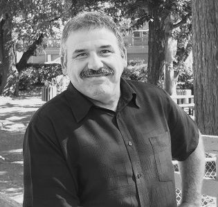 Man with mustache smiles outdoors, wearing a dark shirt; trees in the background.