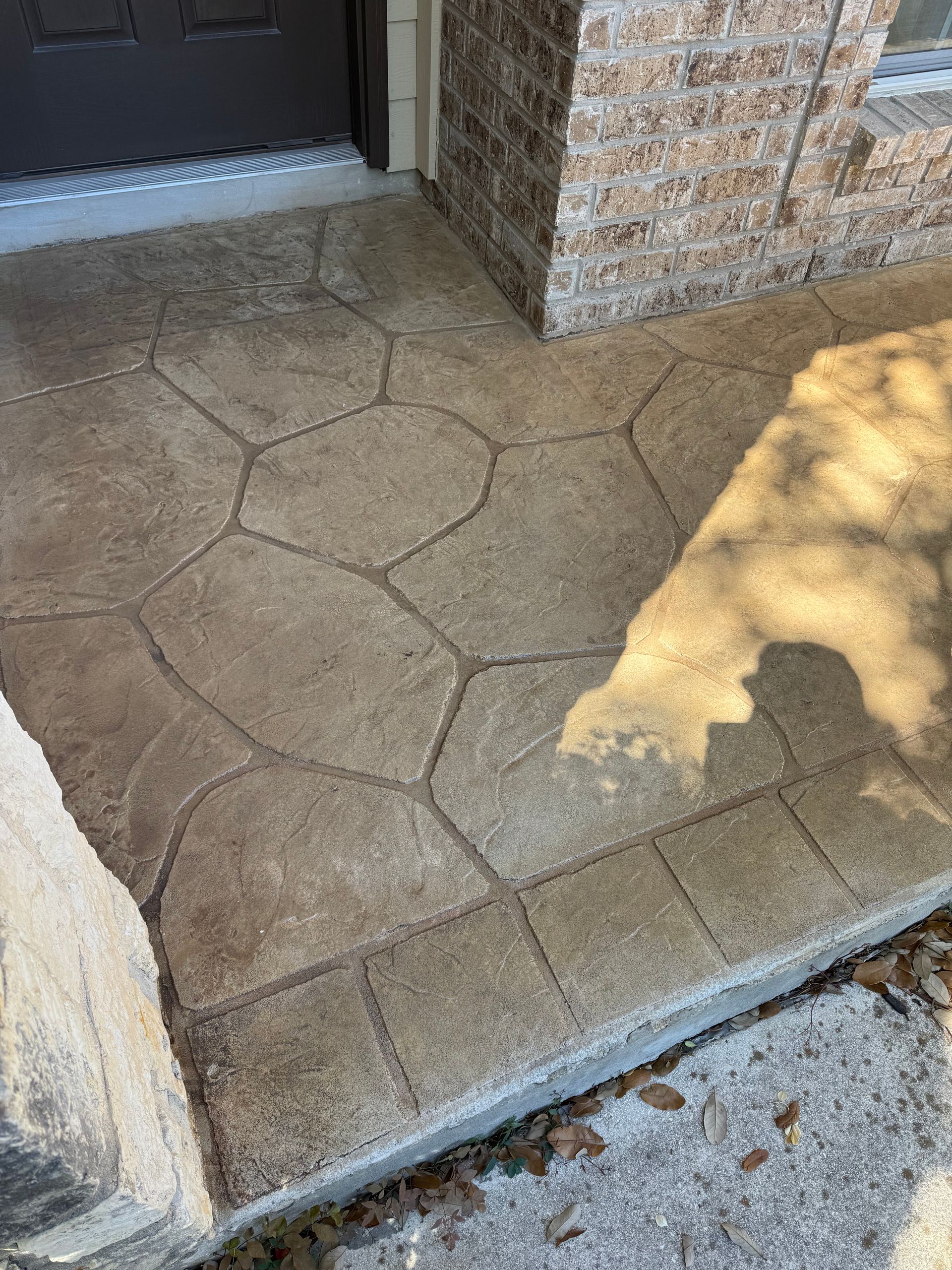 Stamped concrete patio beside a brick wall, with sunlight and a shadow across the ground.