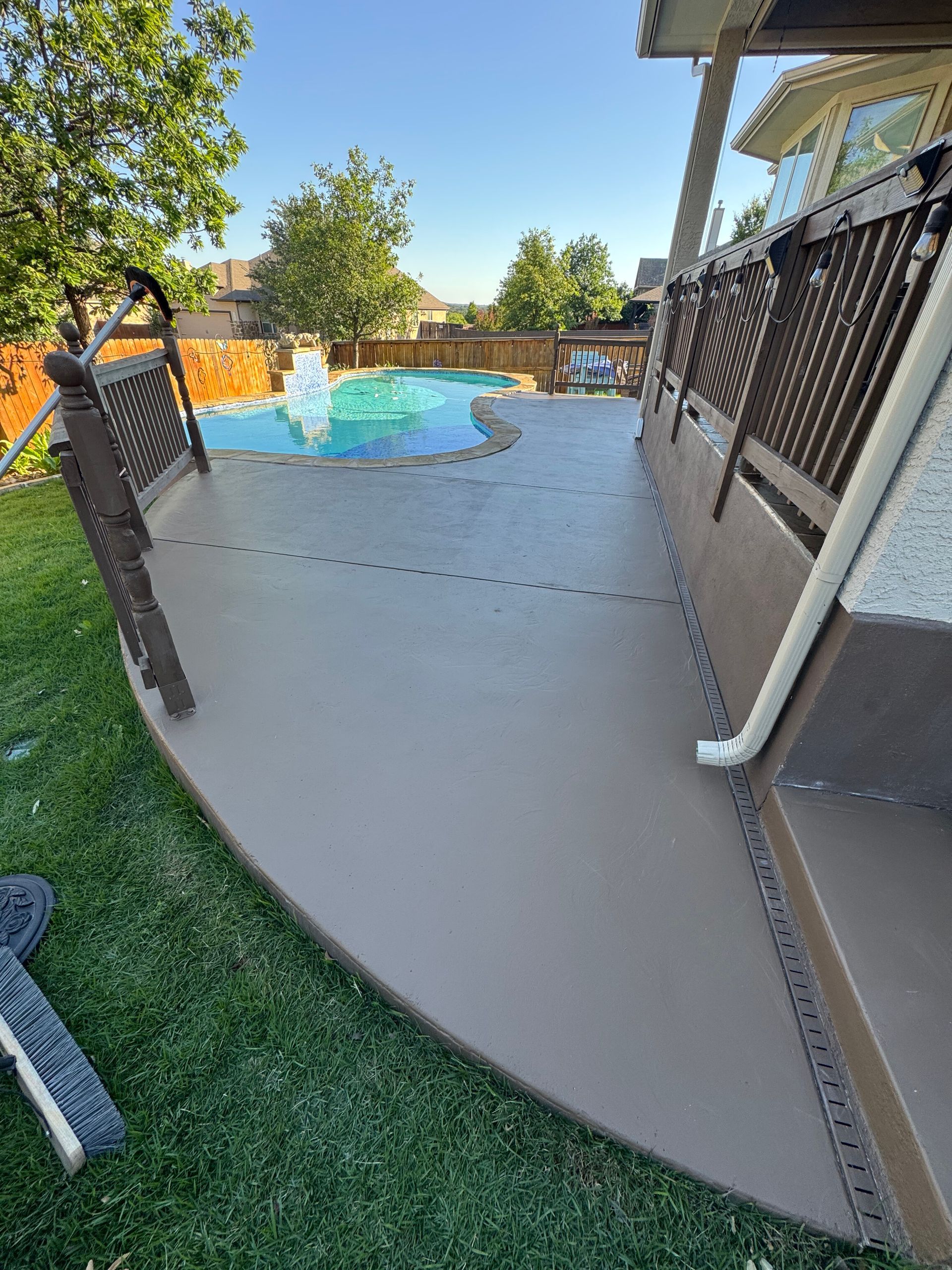 Curved concrete pool deck with black railing, overlooking a backyard pool and trees.