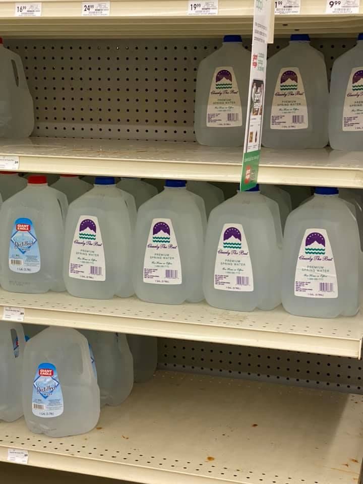 Shelves of bottled water in a store; jugs of various brands with blue and white labels.