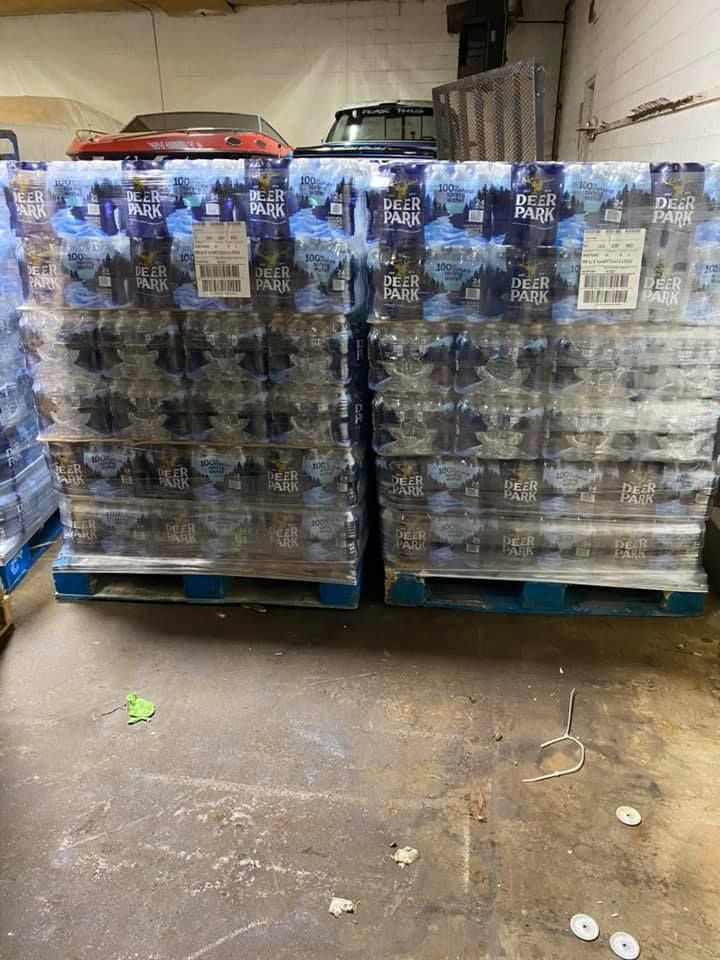Two pallets of bottled water, wrapped in plastic, sit on a concrete floor in a warehouse.