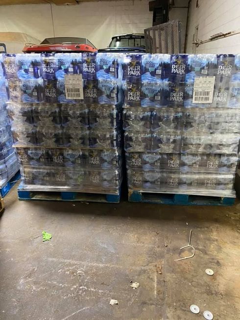 Two pallets of bottled water, wrapped in plastic, sit on a concrete floor in a warehouse.