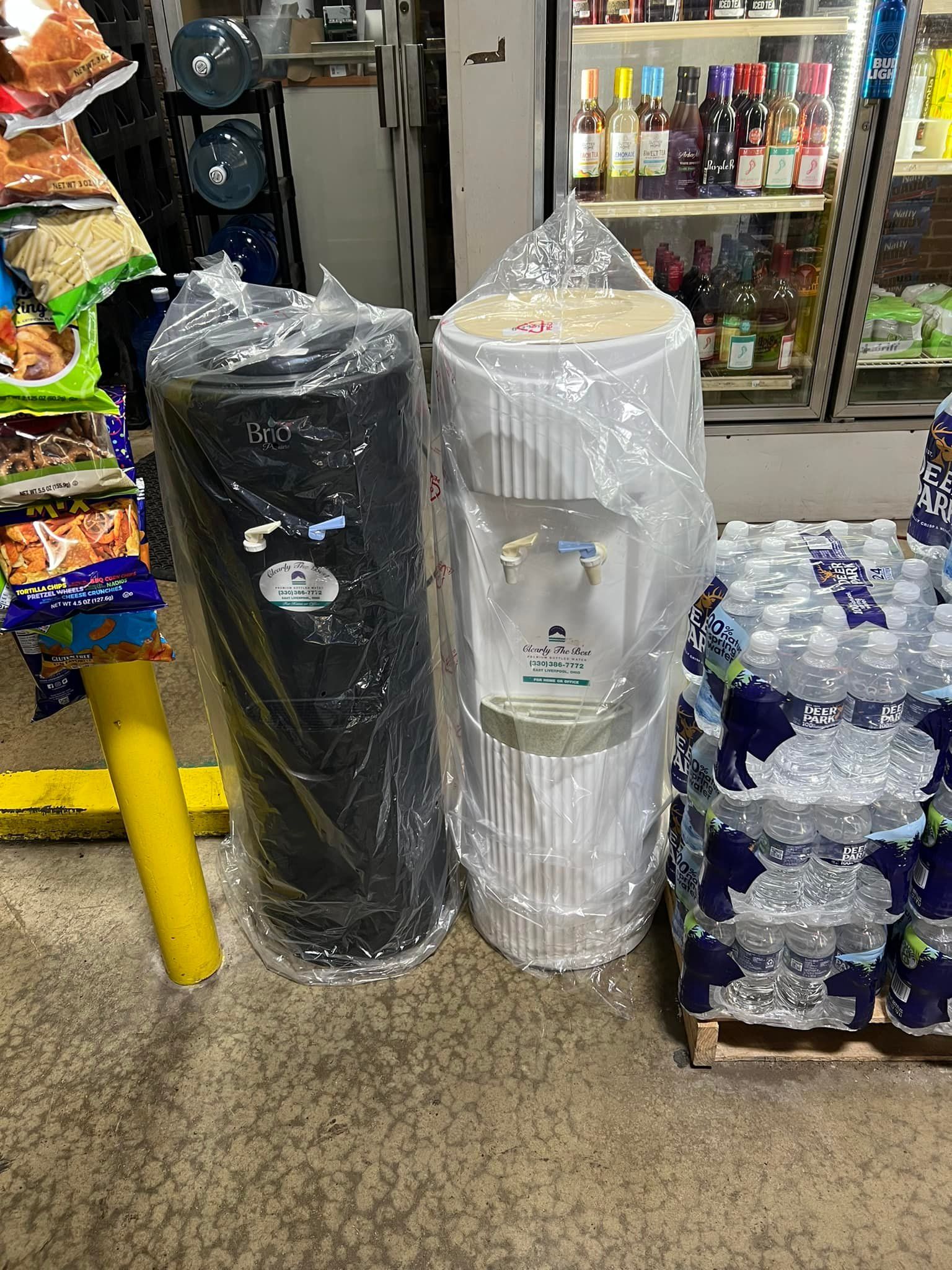 Two water cooler tanks, one black and one white, wrapped in plastic, next to bottled water, in a store.