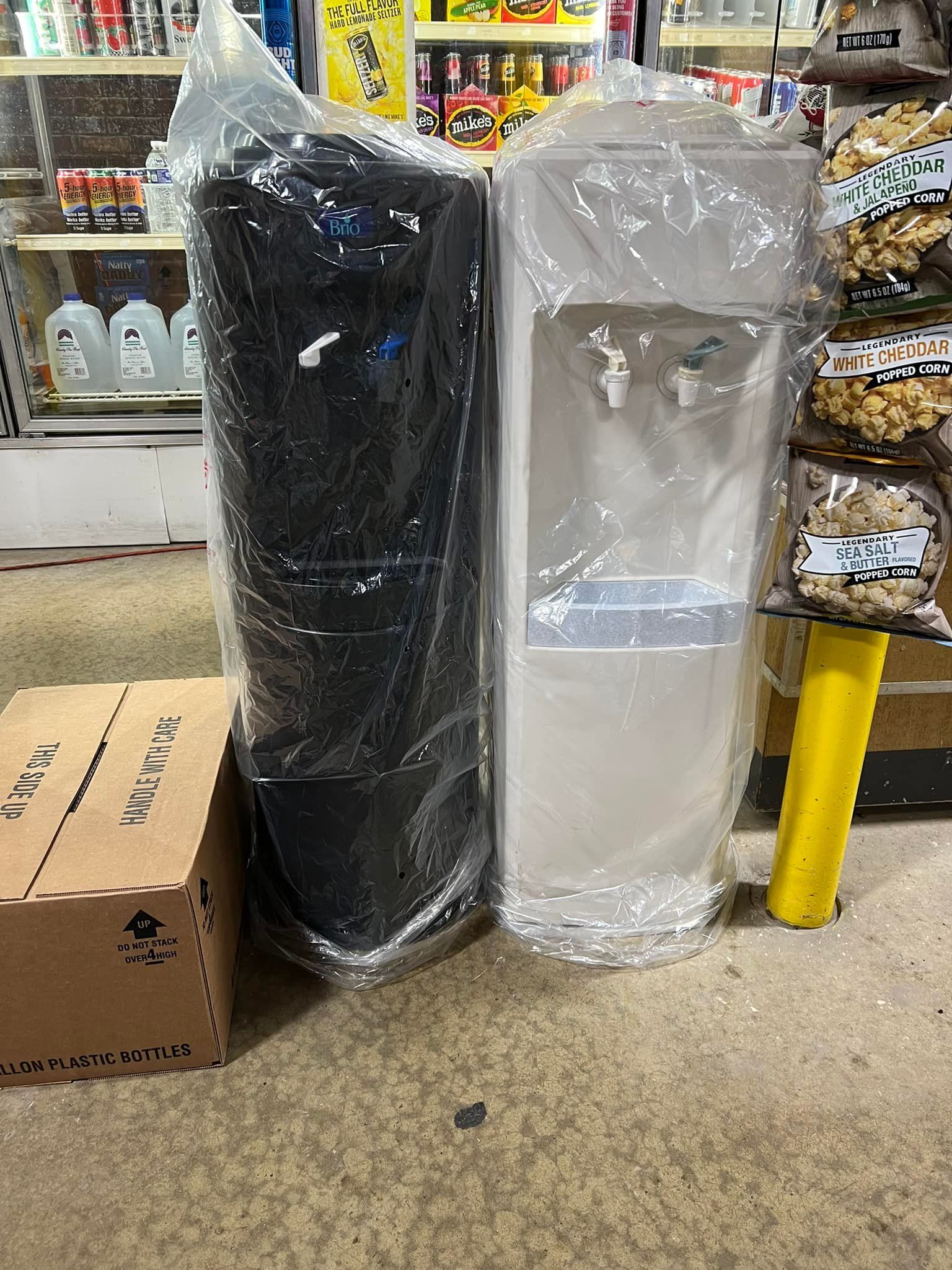 Two new water coolers, one black and one white, still in their plastic wrap, are in a store setting.