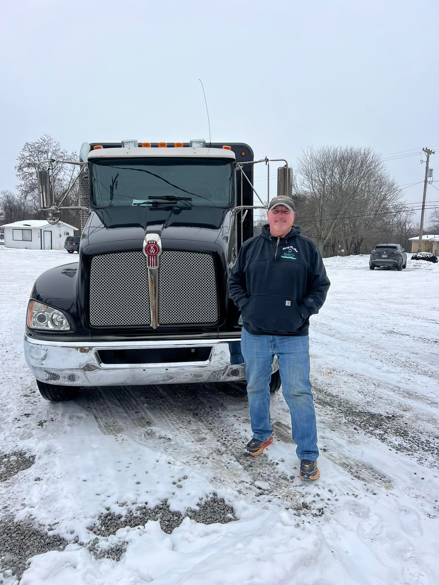 Man standing in front of a black semi-truck in a snowy area. He wears a dark hoodie, jeans, and a cap.