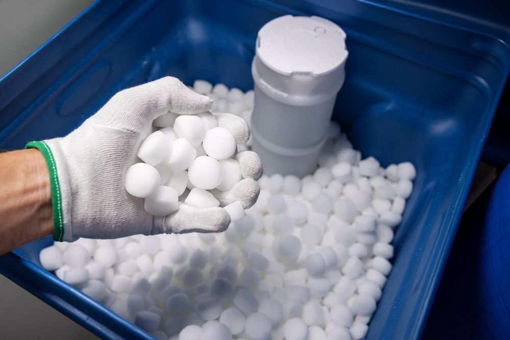 Person holding a handful of white, spherical objects, possibly salt pellets, over a blue container.
