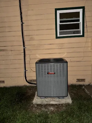 AC unit outside a building with a window, against a tan wall, and a black tube running down the side.
