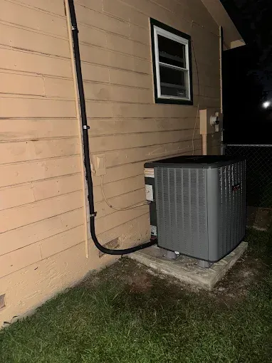 Air conditioning unit outside a beige building next to a window and electrical conduit at night.