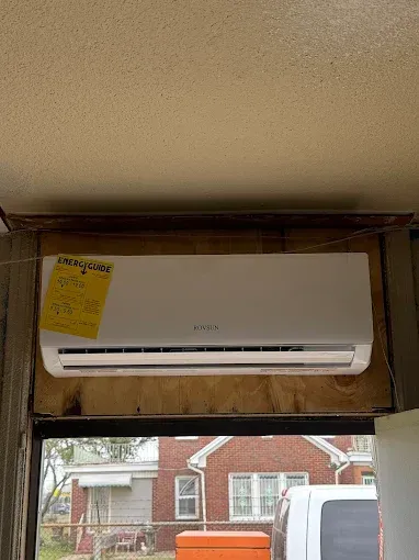 White air conditioner unit mounted on plywood above a window, with a yellow sticky note.