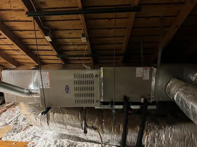 HVAC unit installed in an attic; silver rectangular unit with attached ductwork, exposed rafters and insulation.