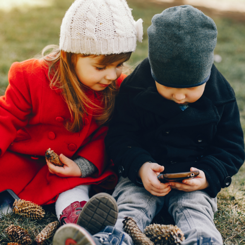 A boy and a girl are sitting on the grass looking at a cell phone