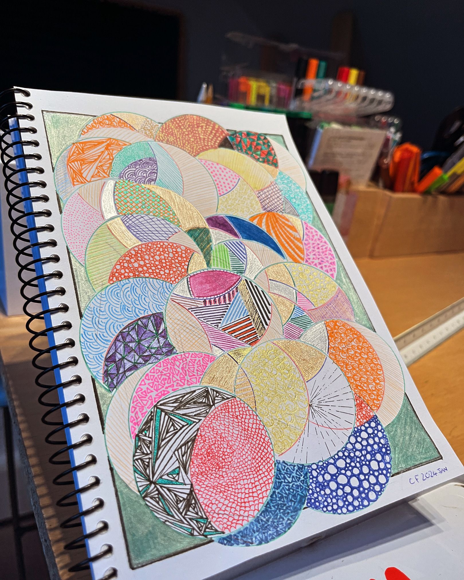 Spiral-bound sketchbook with a colorful, patterned drawing of overlapping circles, set on a wooden surface.
