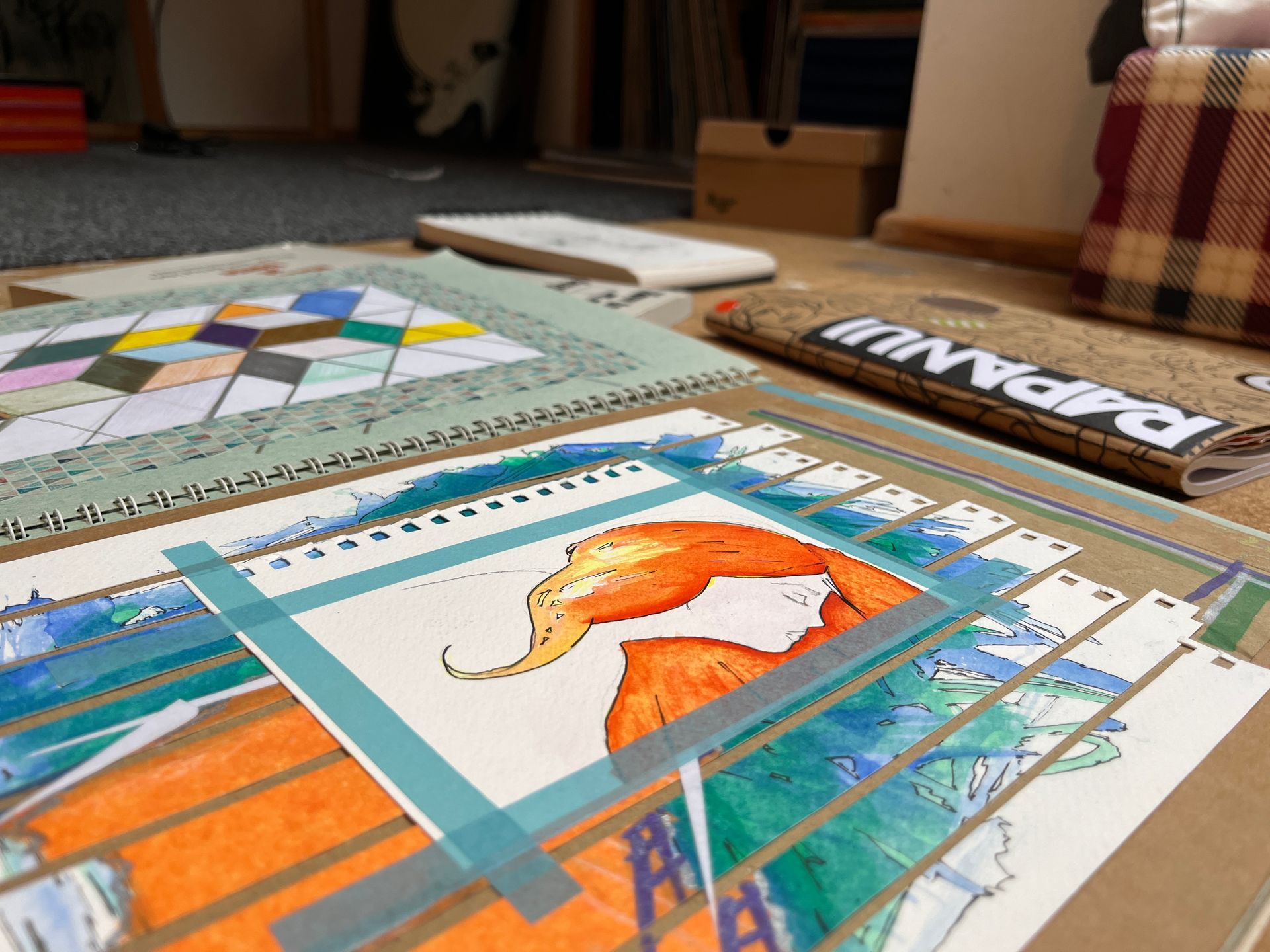 Artwork in progress; close-up of a watercolour painting, taped to a sketchbook on a table, with other art supplies.