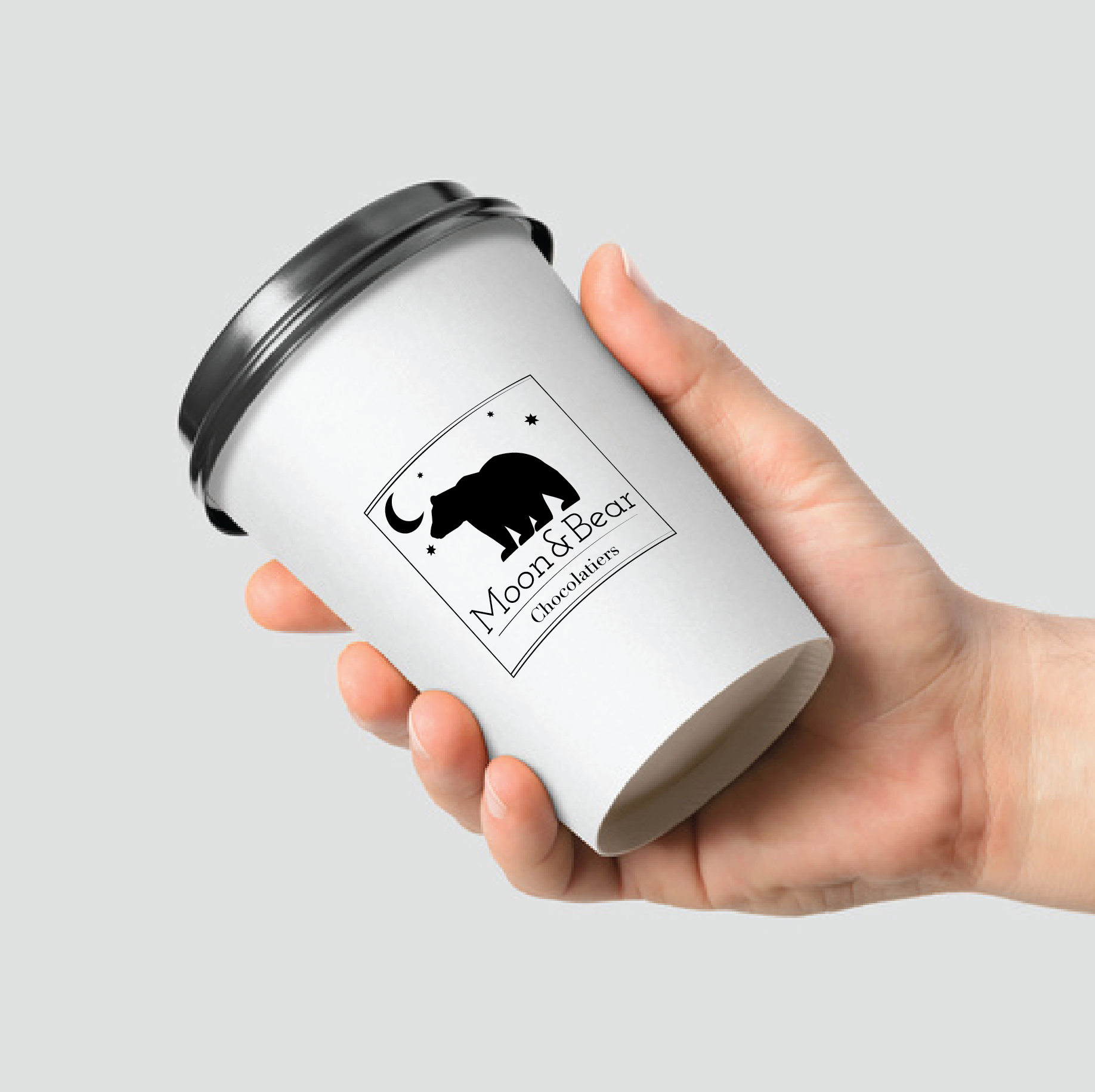 Hand holding a white coffee cup with black lid, Moon & Bear logo on it.
