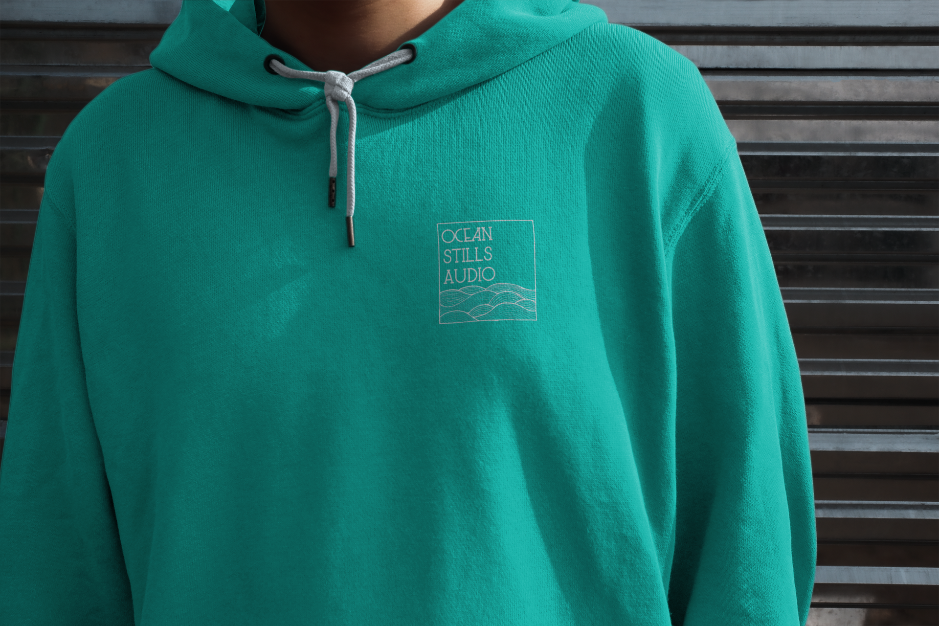Teal hoodie with white logo on the chest, drawstring hood. Person in front of a corrugated metal wall.