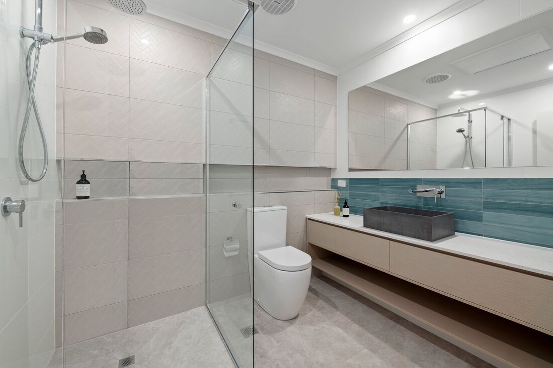 A modern bathroom featuring a glass-enclosed shower, a white toilet, and a vanity with a dark basin against teal tiles. — Foreshore Kitchens in Coolum Beach, QLD