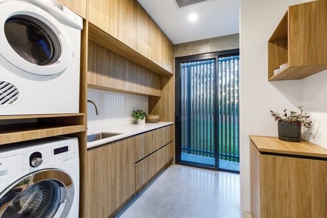 A modern laundry room with wood cabinets, white countertops, stacked washer and dryer, and a glass door leading outside. — Foreshore Kitchens in Coolum Beach, QLD