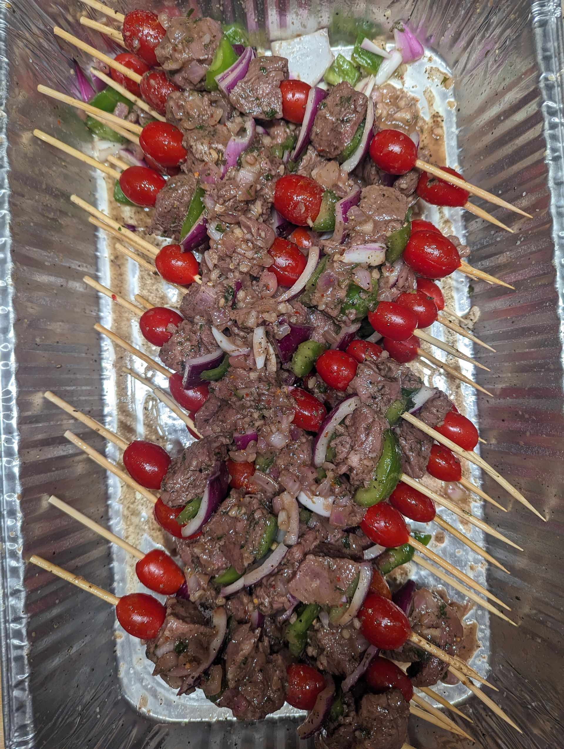 A tray of food with meat , tomatoes , onions and peppers on skewers.