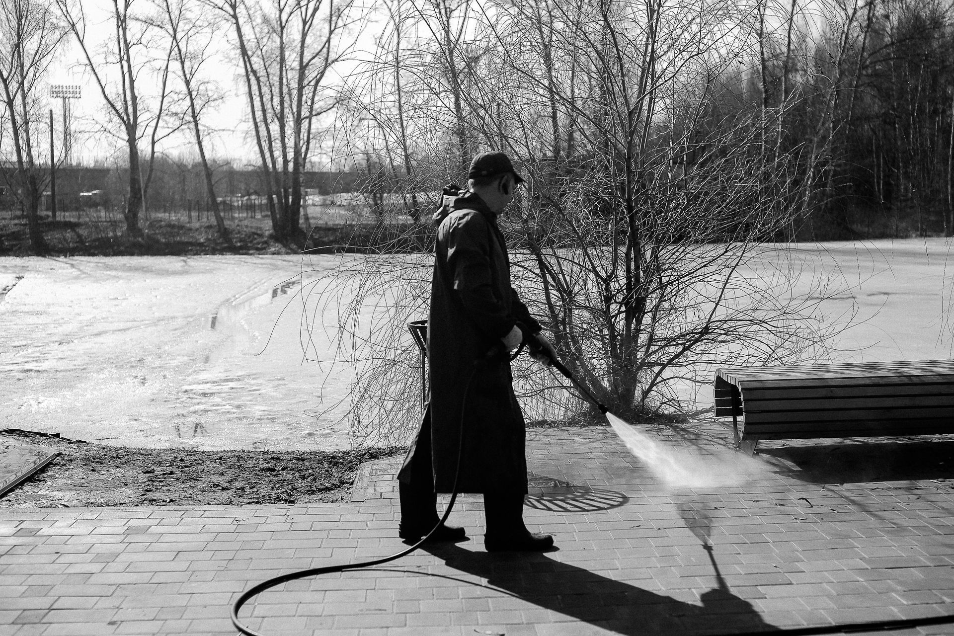 Person in a long coat power washing a brick patio; trees and a body of water in the background.