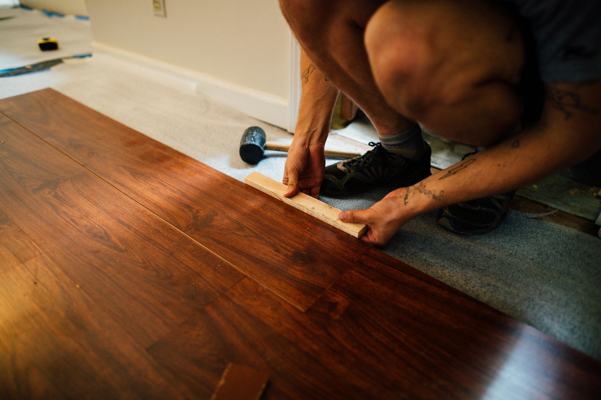 Person installing hardwood flooring, measuring a plank with a ruler, near a wall and a mallet.