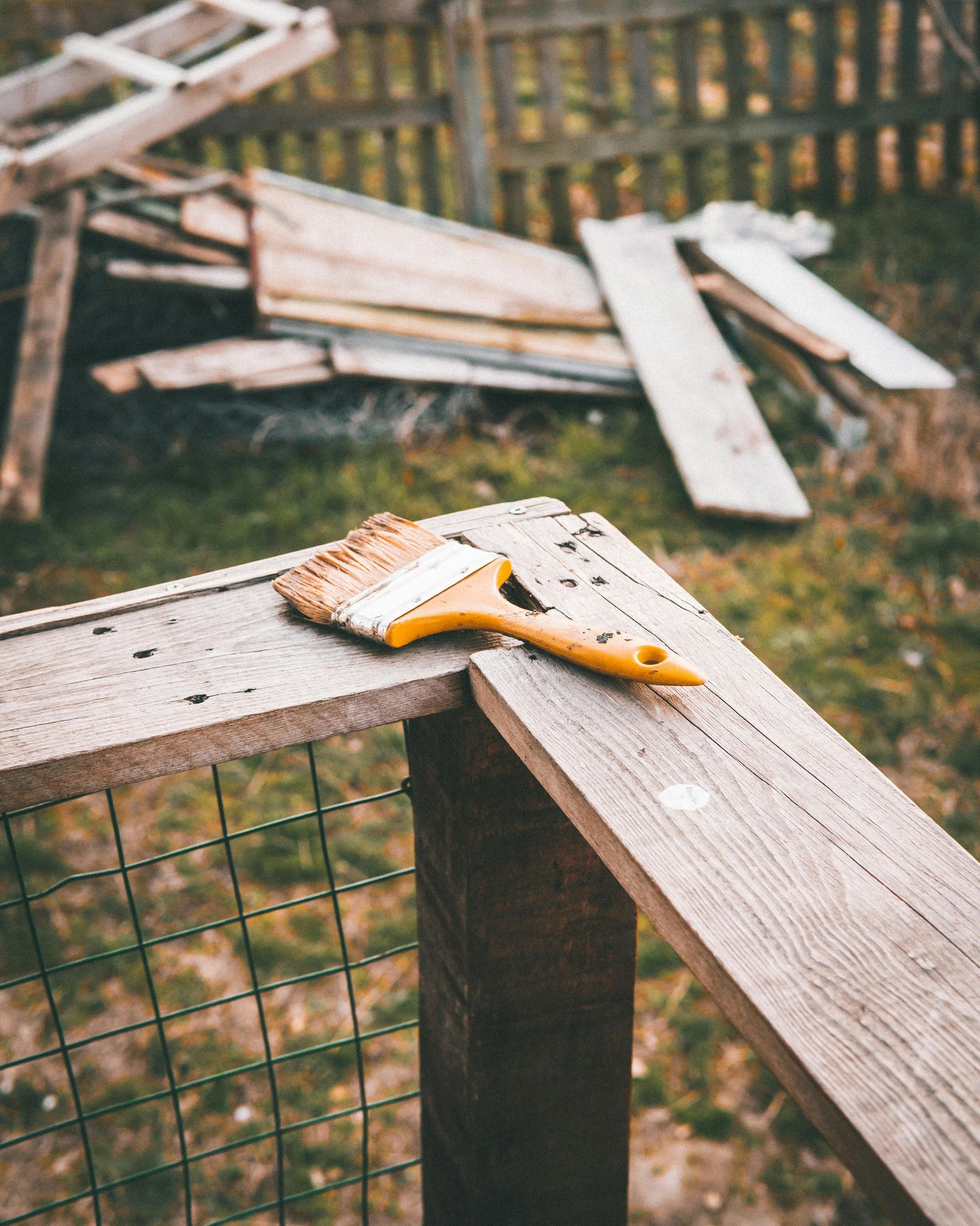 Paintbrush on wooden frame, with scattered planks and fence in background.