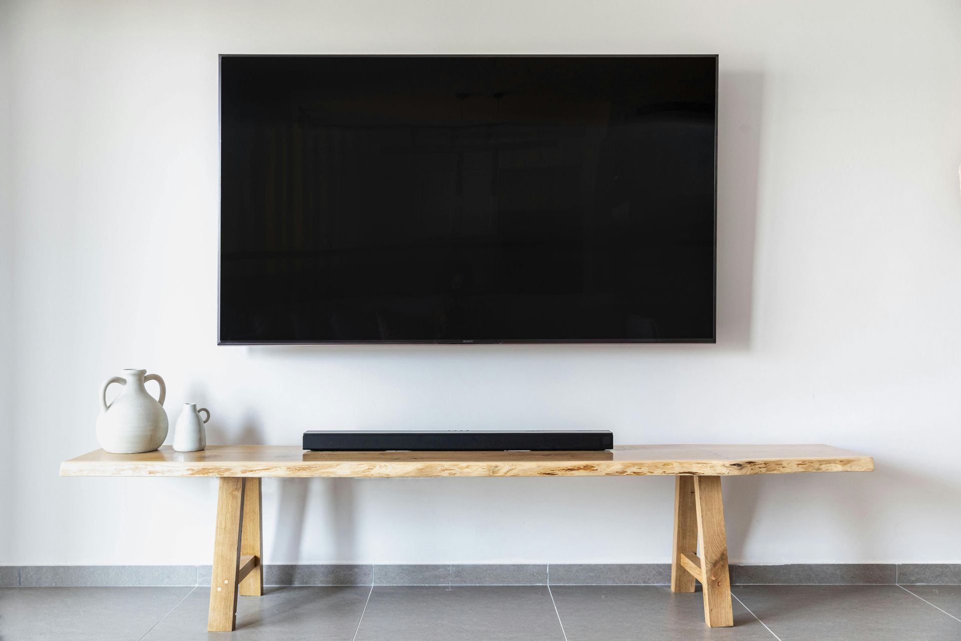 Large black television mounted on white wall, above a wooden bench with soundbar and decorative vases.