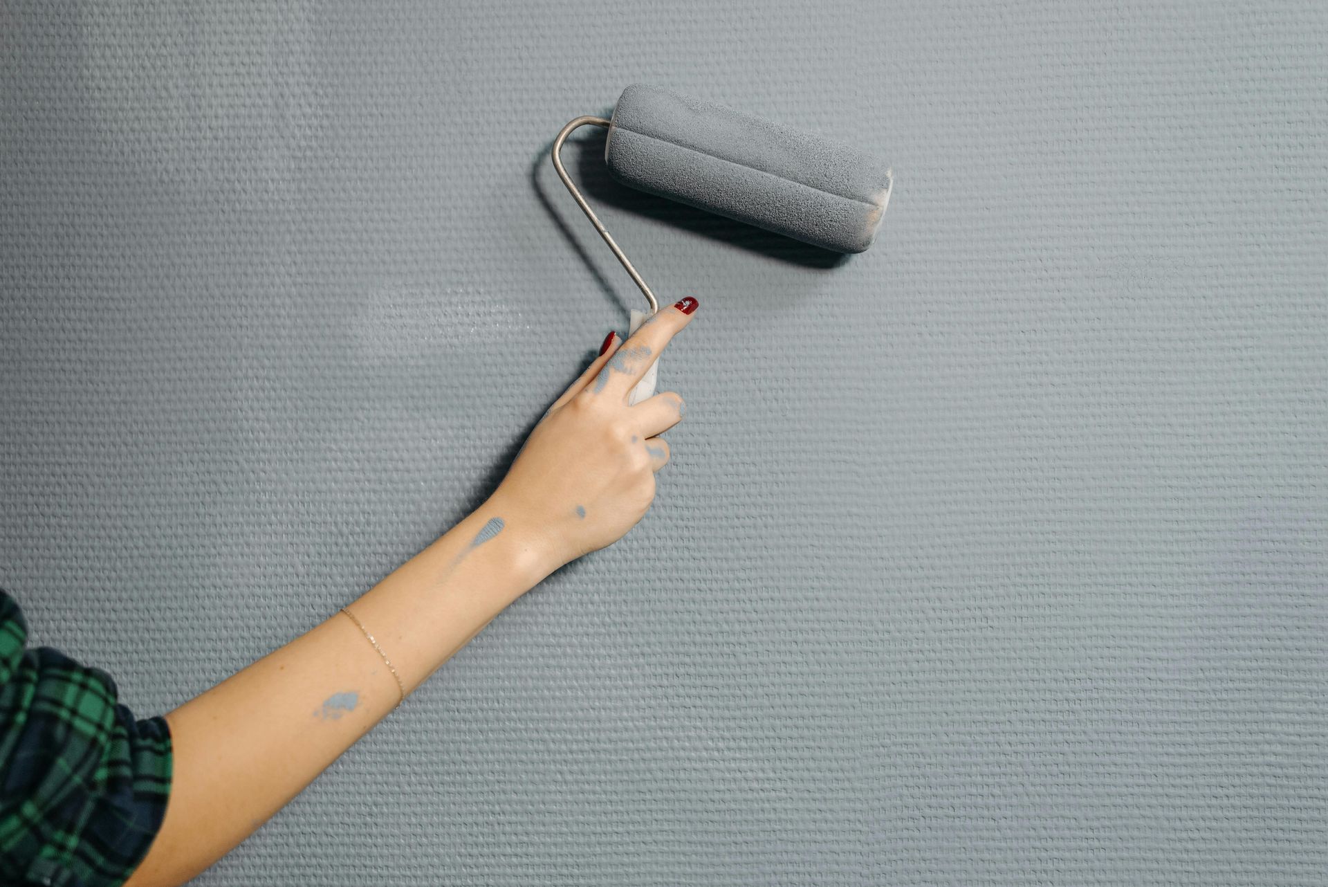 Hand with red nails using a paint roller on a textured gray wall, wearing a plaid shirt.