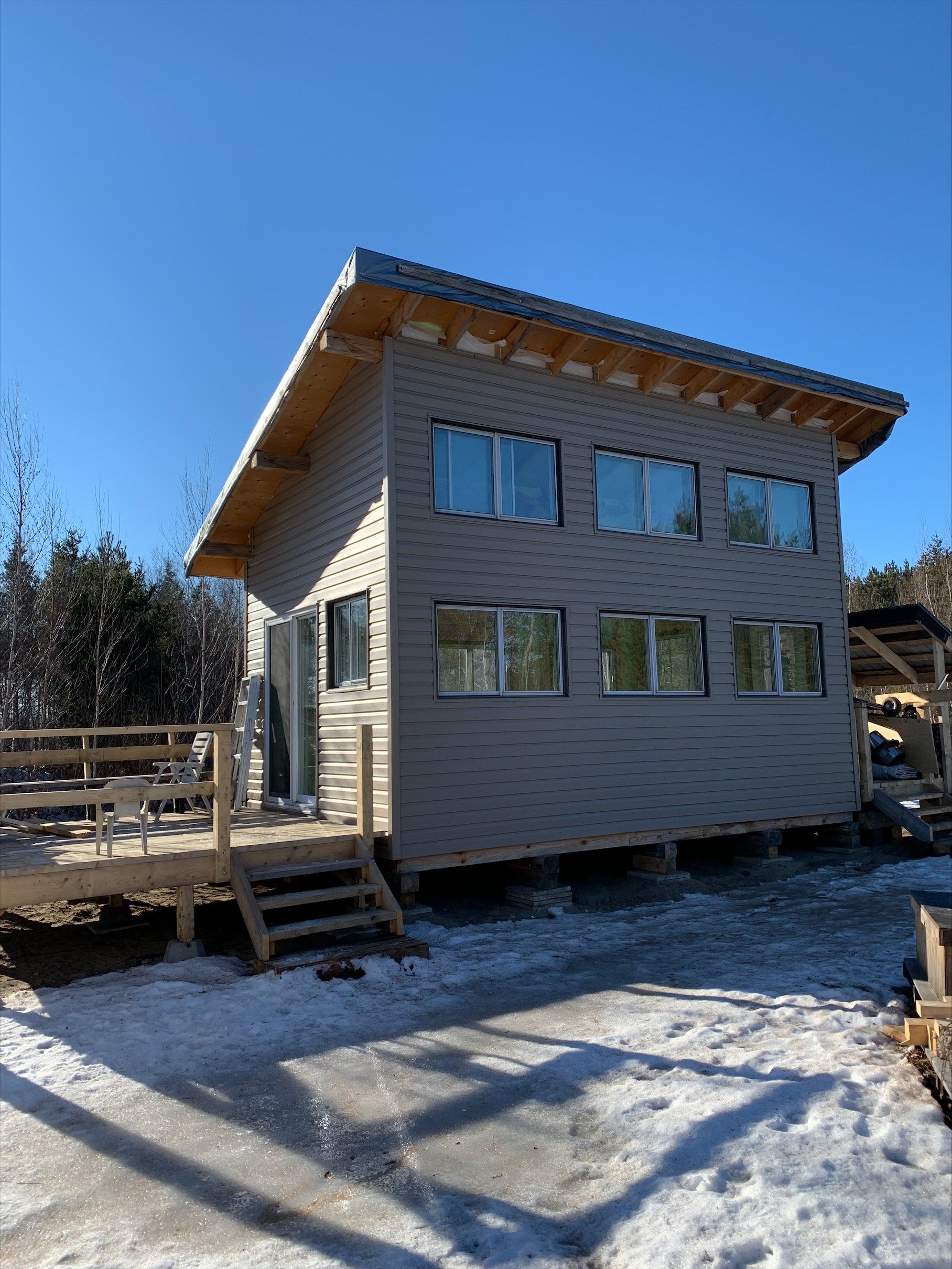 Maison à deux étages avec revêtement gris, terrasse en bois et sol enneigé par une journée ensoleillée.