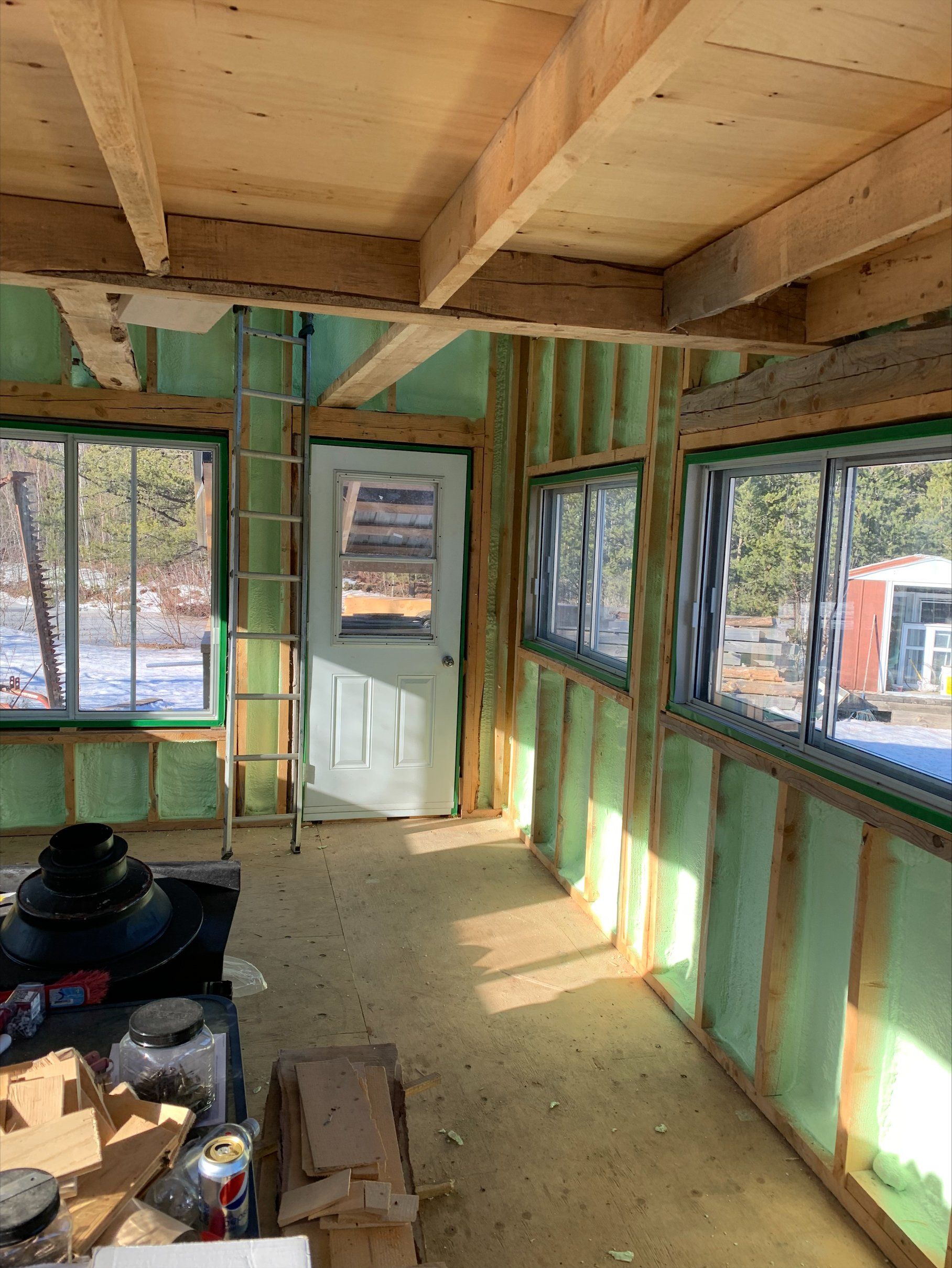 Intérieur d'une petite pièce inachevée avec fenêtres et porte, isolation verte et une échelle appuyée contre un mur.