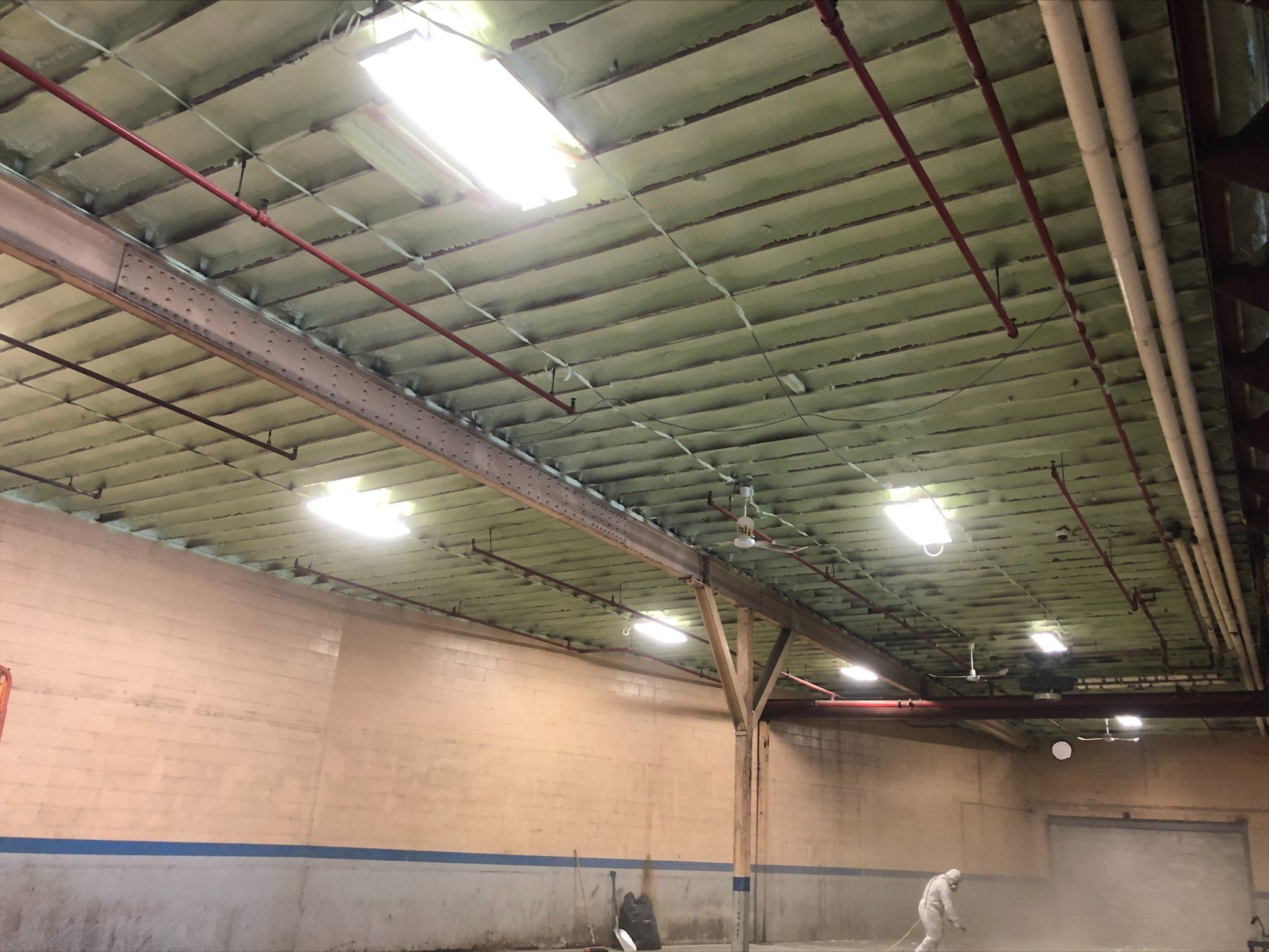 À l'intérieur d'un entrepôt : plafond éclairé par des néons. Des ouvriers balayent. Murs beiges, poutres en bois.