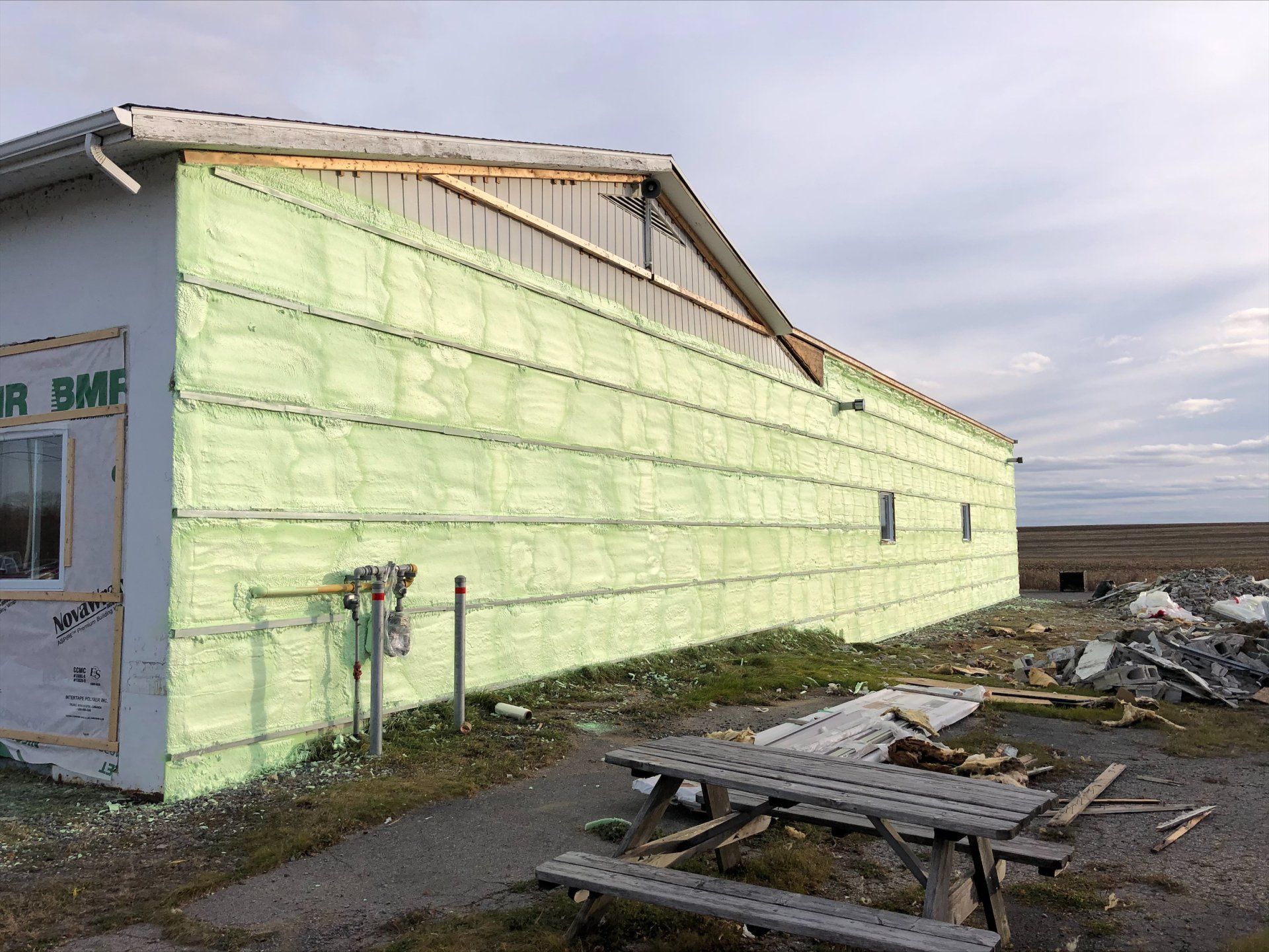 Extérieur du bâtiment en construction; panneaux isolants vert clair, toiture non finie, table de pique-nique devant.