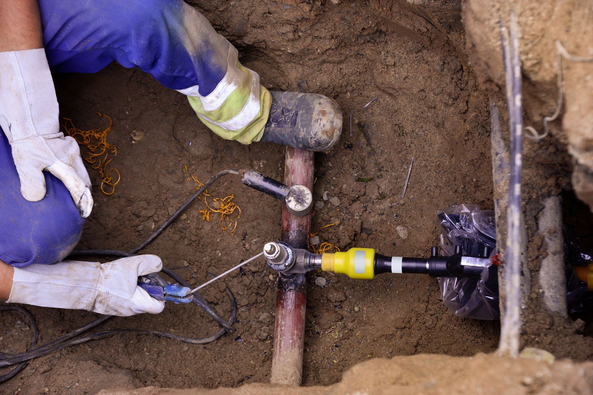 A worker repairs a pipe underground, using tools and wearing gloves and a blue jumpsuit.
