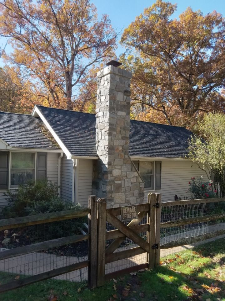 Stone chimney beside a gray house, with a wooden gate and autumn trees in the yard