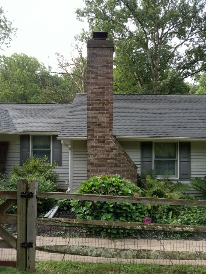 Single-story house with gray siding and a tall brick chimney behind a wooden fence and green shrubs.