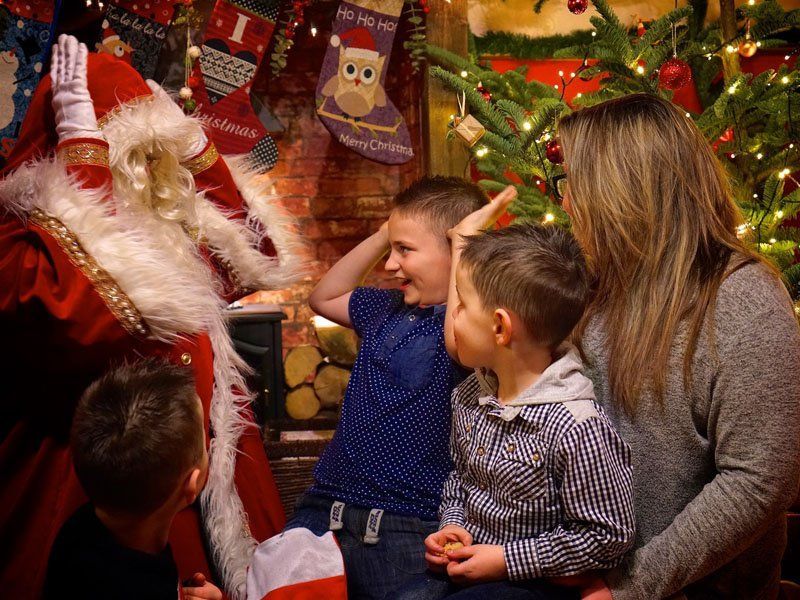 Santa Claus with children, a woman, and Christmas decorations indoors. Children look surprised.