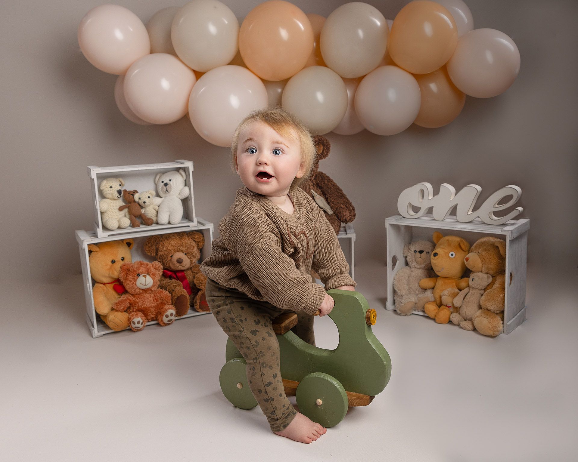 A baby is sitting on a rocking horse in front of teddy bears and balloons.