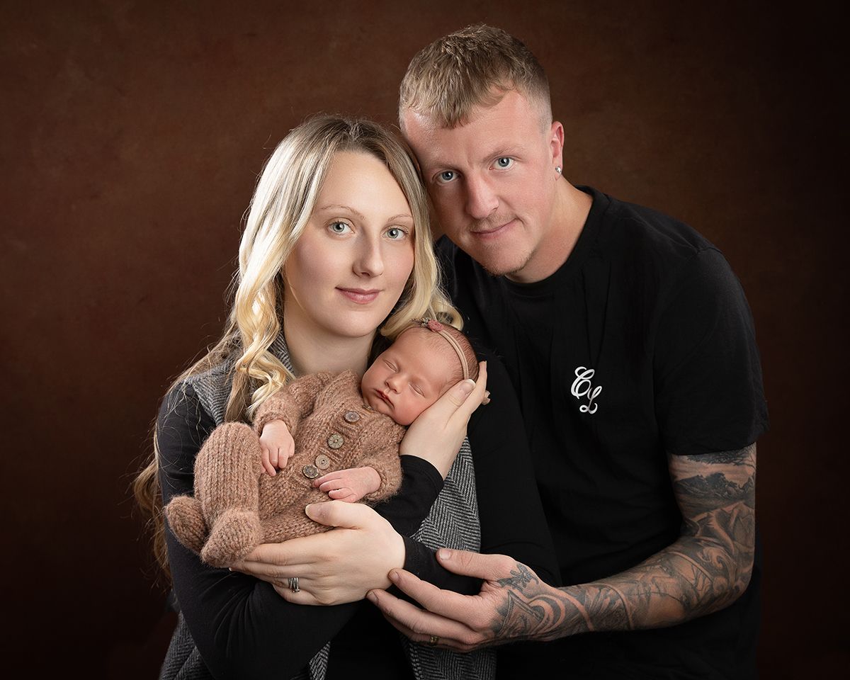 Parents holding a newborn baby, posing for a studio portrait. Brown background.