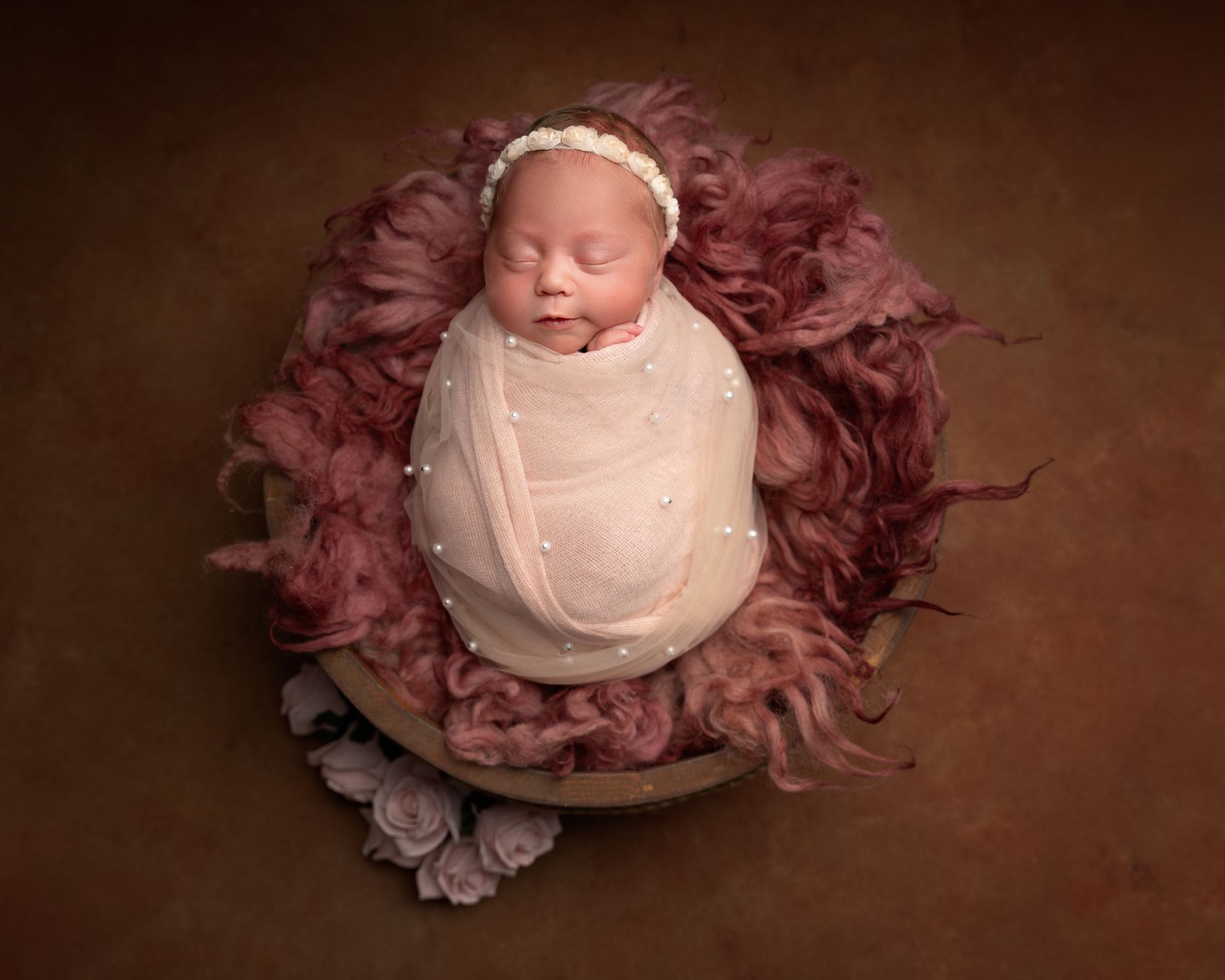 Newborn swaddled in pink blanket with pearls, sleeping in a bowl with fur and headband. Brown background.