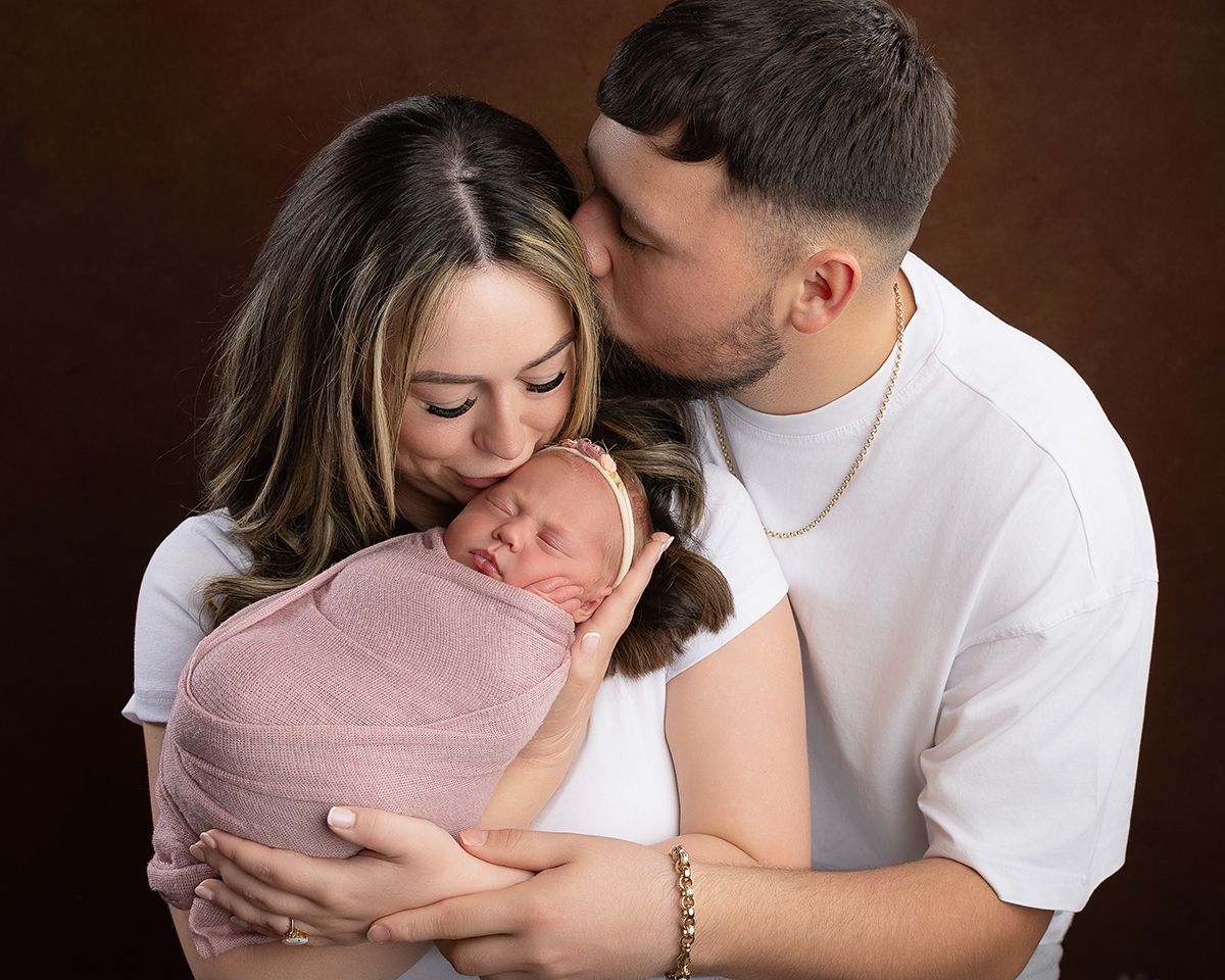 Parents kissing their newborn baby, swaddled in pink, against a brown backdrop.