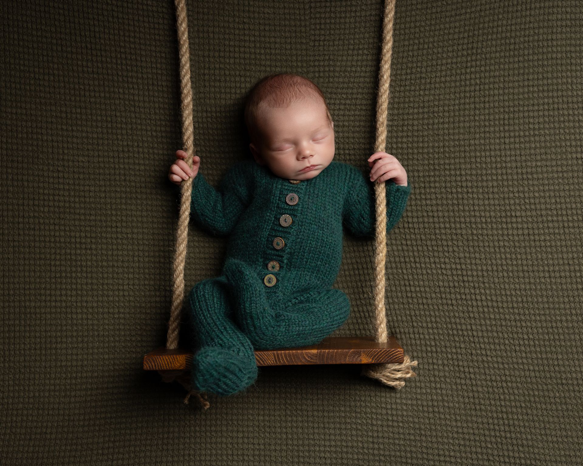 Newborn baby in green onesie asleep on a wooden swing against a dark green backdrop.