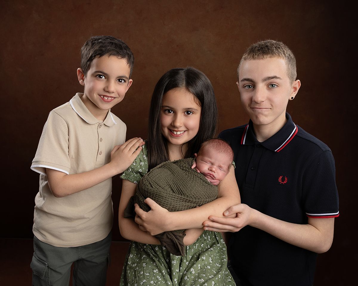 Family portrait: Three siblings with newborn baby; girl holding baby; all smiling, brown background.