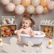 A baby in a bathtub, surrounded by teddy bears, balloons, and duck toys, celebrating their first birthday.
