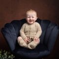 Smiling baby in a fluffy beige outfit sitting in a navy blue velvet chair against a brown backdrop.