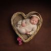 Newborn baby asleep in a heart-shaped wicker basket; wearing a light brown outfit and a headband.