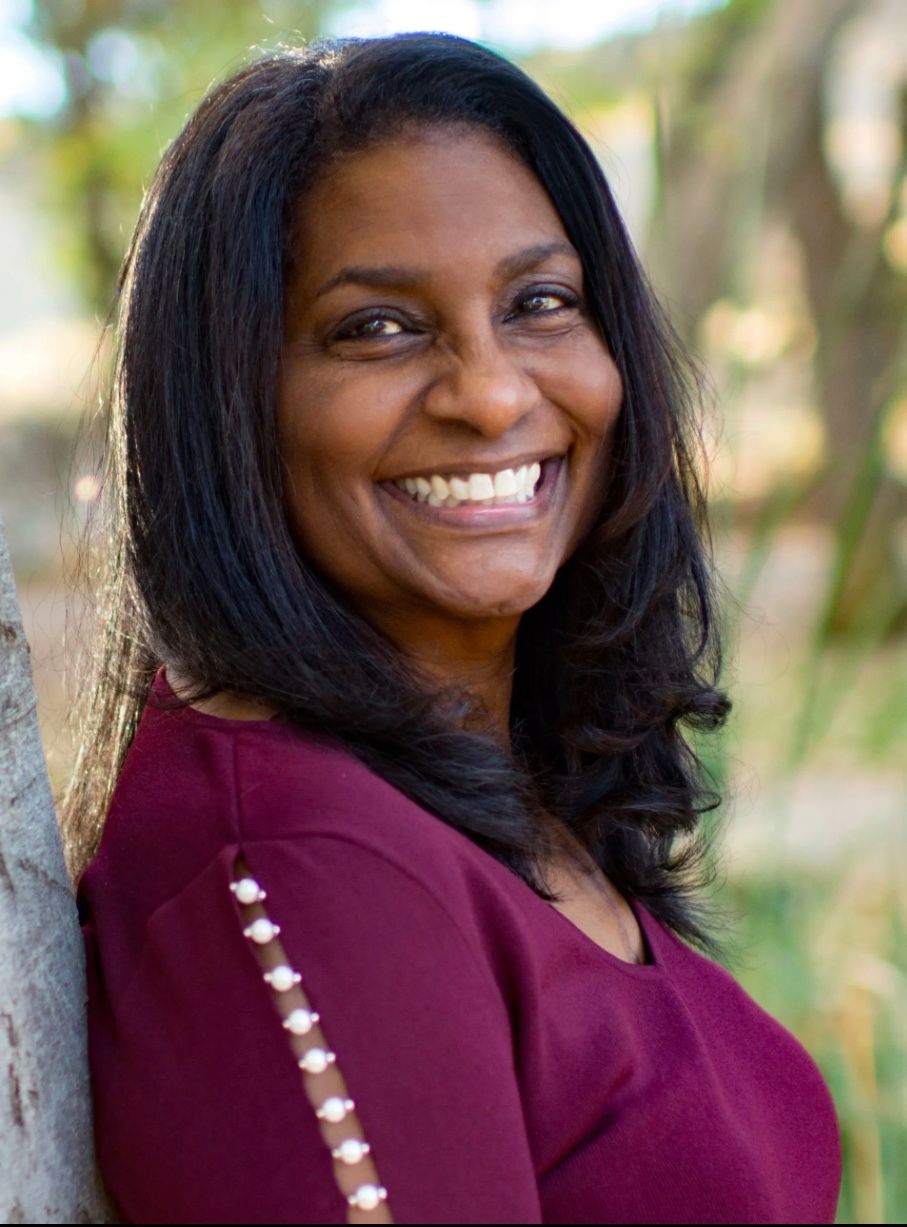 A woman in a red shirt is smiling and leaning against a tree