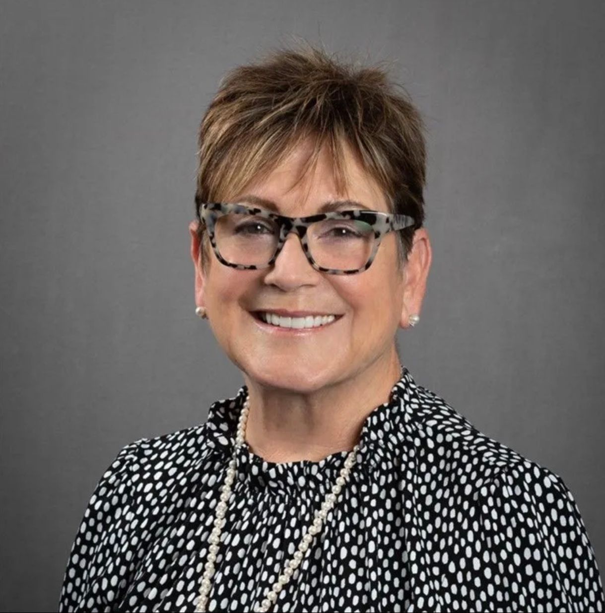 A woman wearing glasses and a pearl necklace is smiling for the camera.